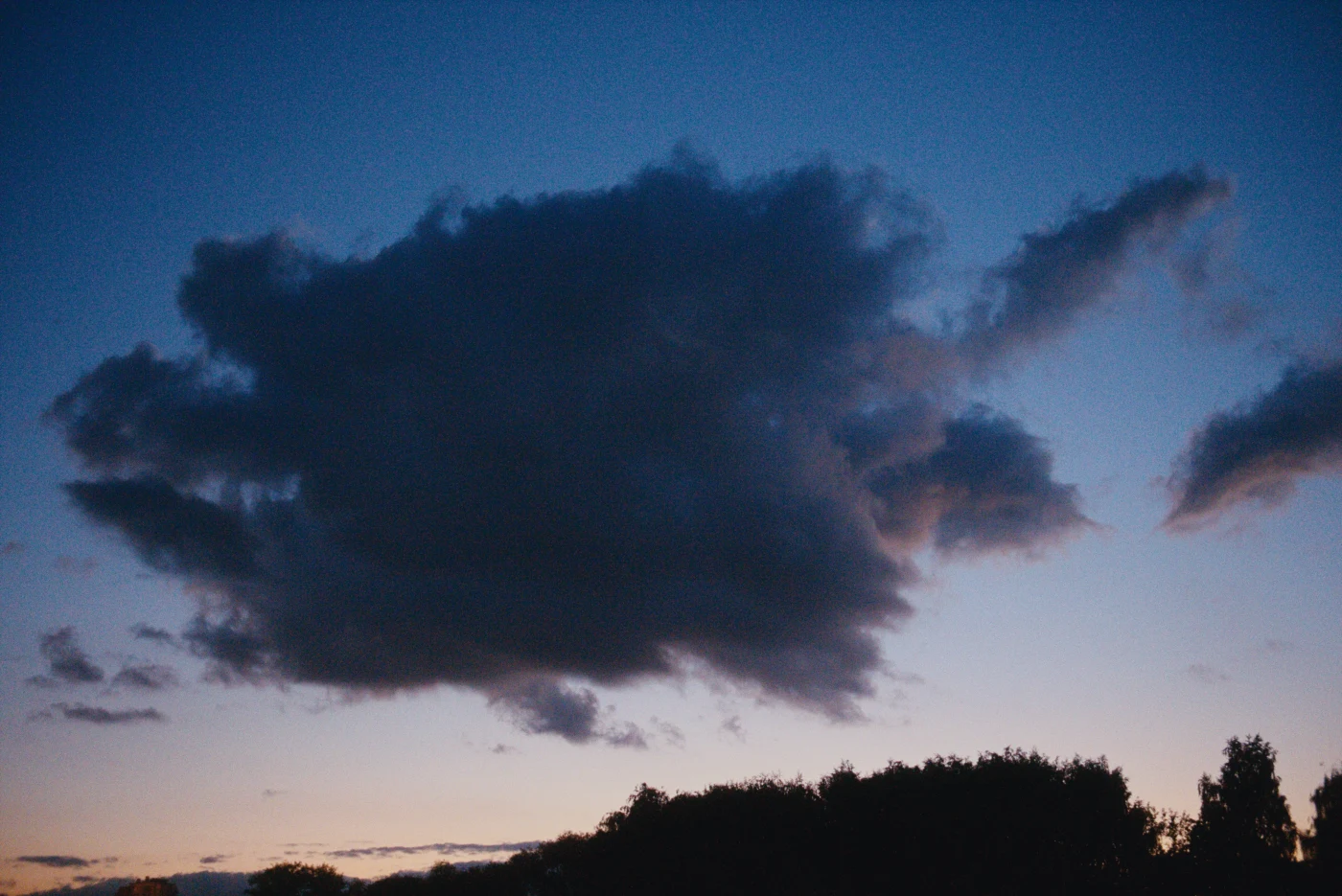 A dark cloud against the evening sky, with lighter clouds and silhouettes of trees below.