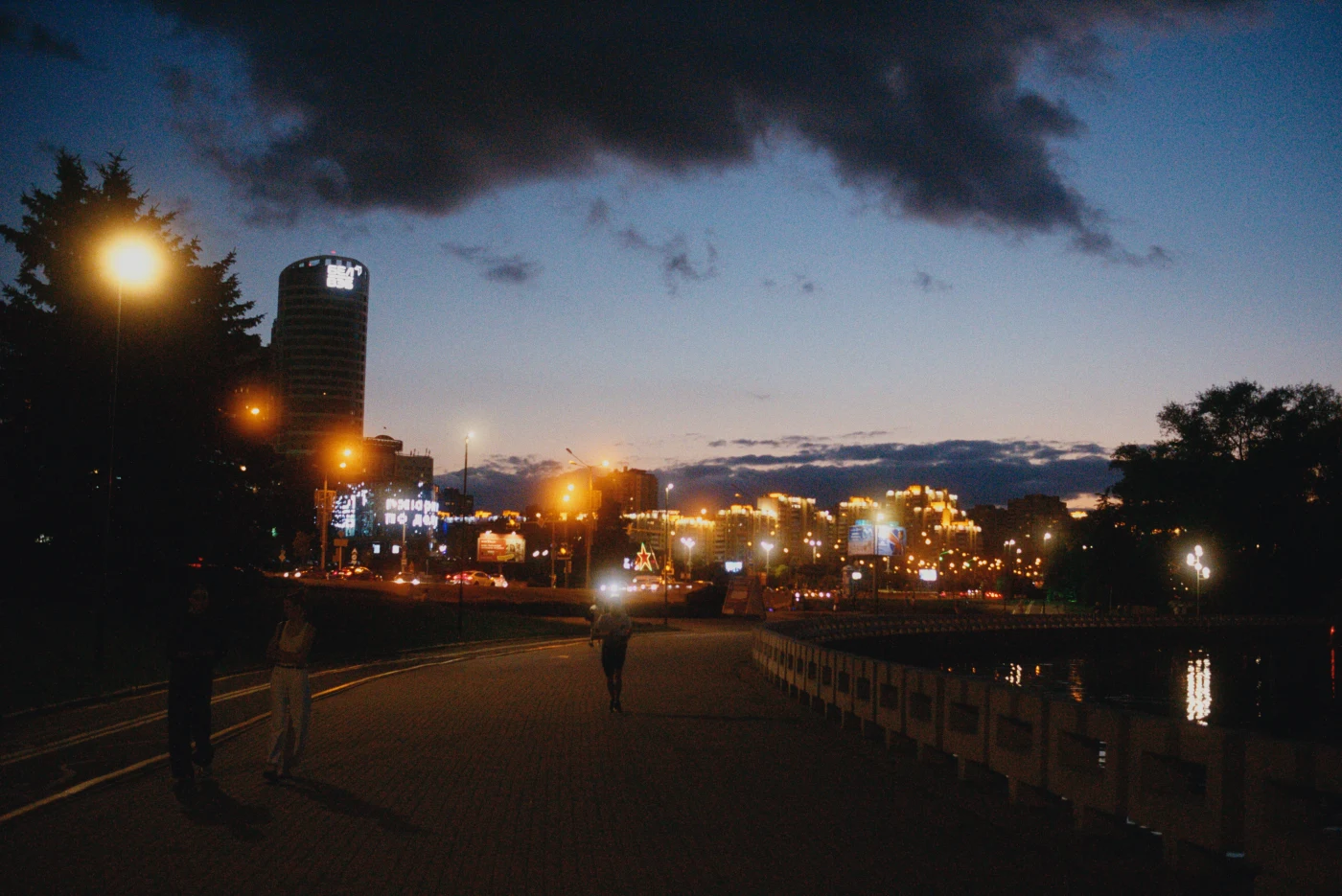 A nighttime cityscape with an illuminated waterfront and silhouettes of people walking.