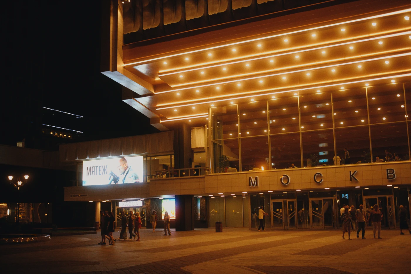 A nighttime view of a brightly illuminated building with a sign reading “МОСКВА”, with people near the entrance.