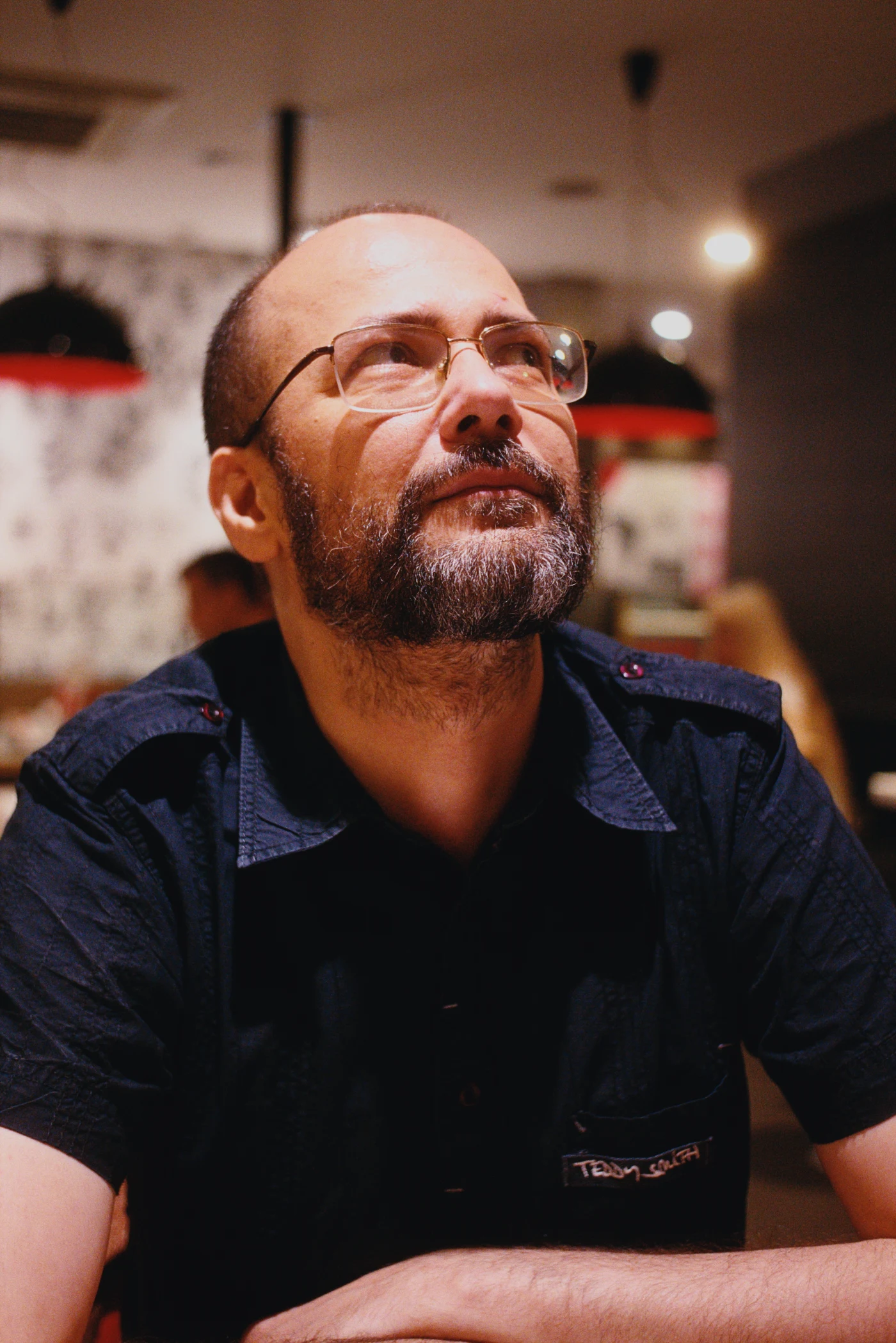 A bearded man wearing glasses looks upward while sitting at a table in a café. Blurred figures and the café interior are visible in the background.