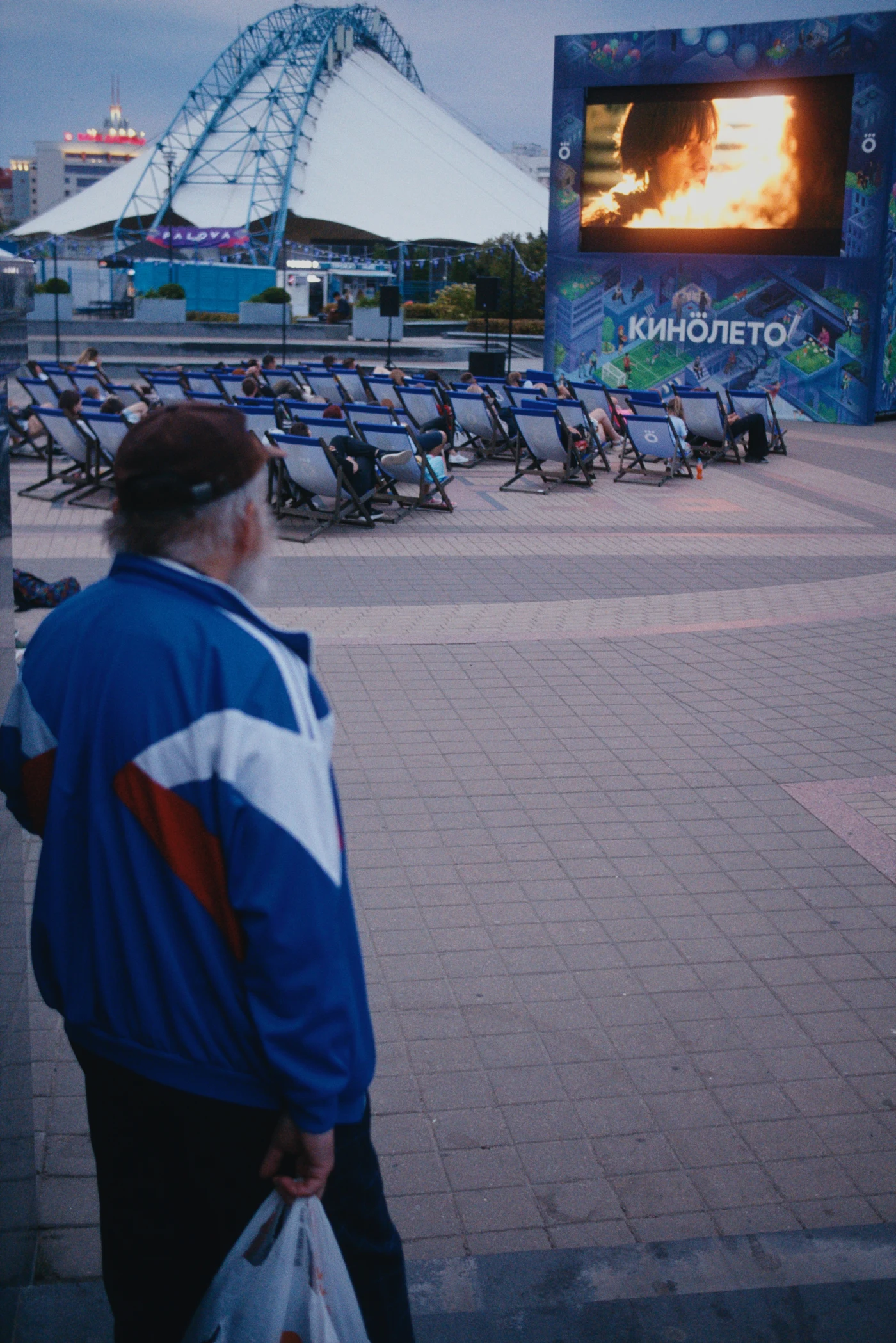 Человек в спортивной куртке смотрит на экран с фильмом на открытом кинотеатре. На заднем плане — крыша павильона.