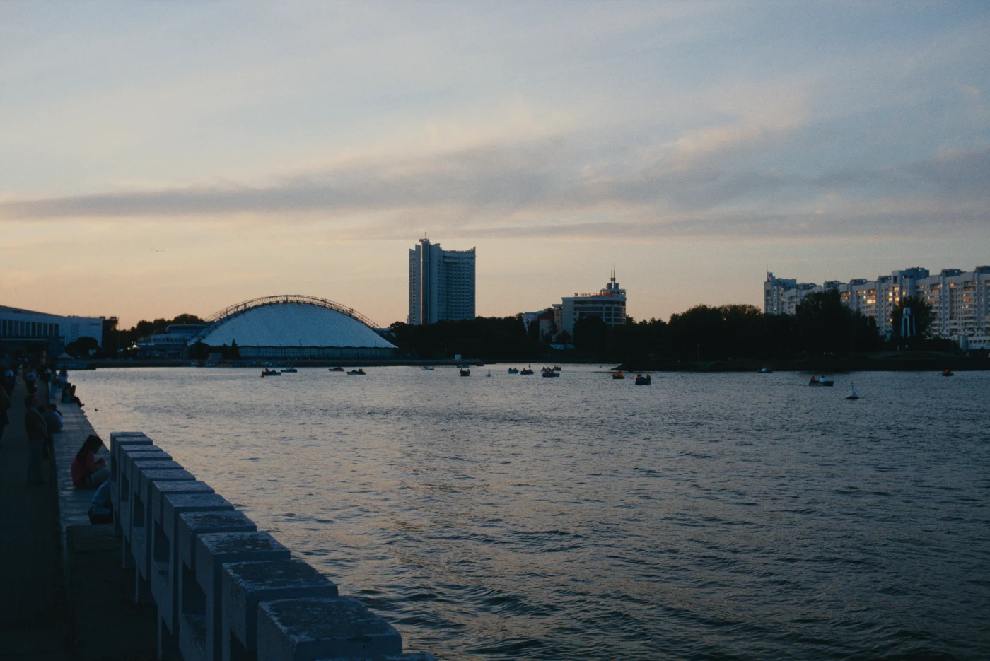 Вечерний пейзаж реки с лодками, на фоне зданий и арки моста. Небо окрашено в мягкие оттенки заката.
