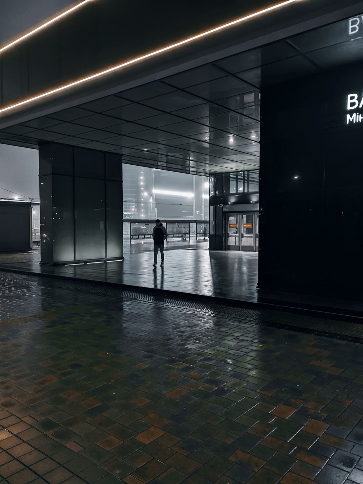 Человек стоит под светом в темном городском пространстве, отражения на мокром асфальте.