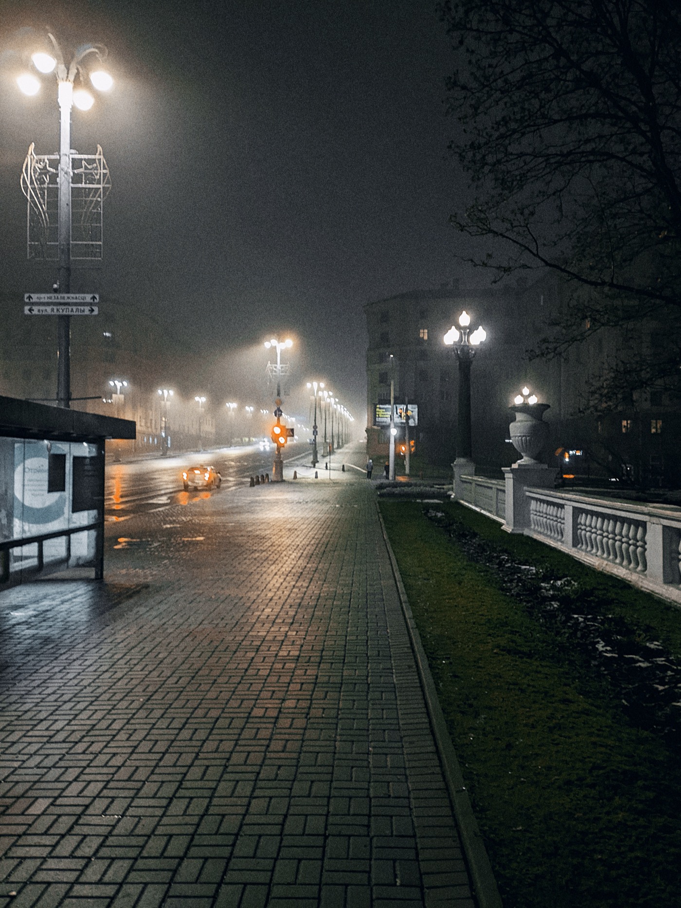 Туманная ночь на улице, освещенной фонарями. Пустая дорога и тротуар, видны огни автомобилей.