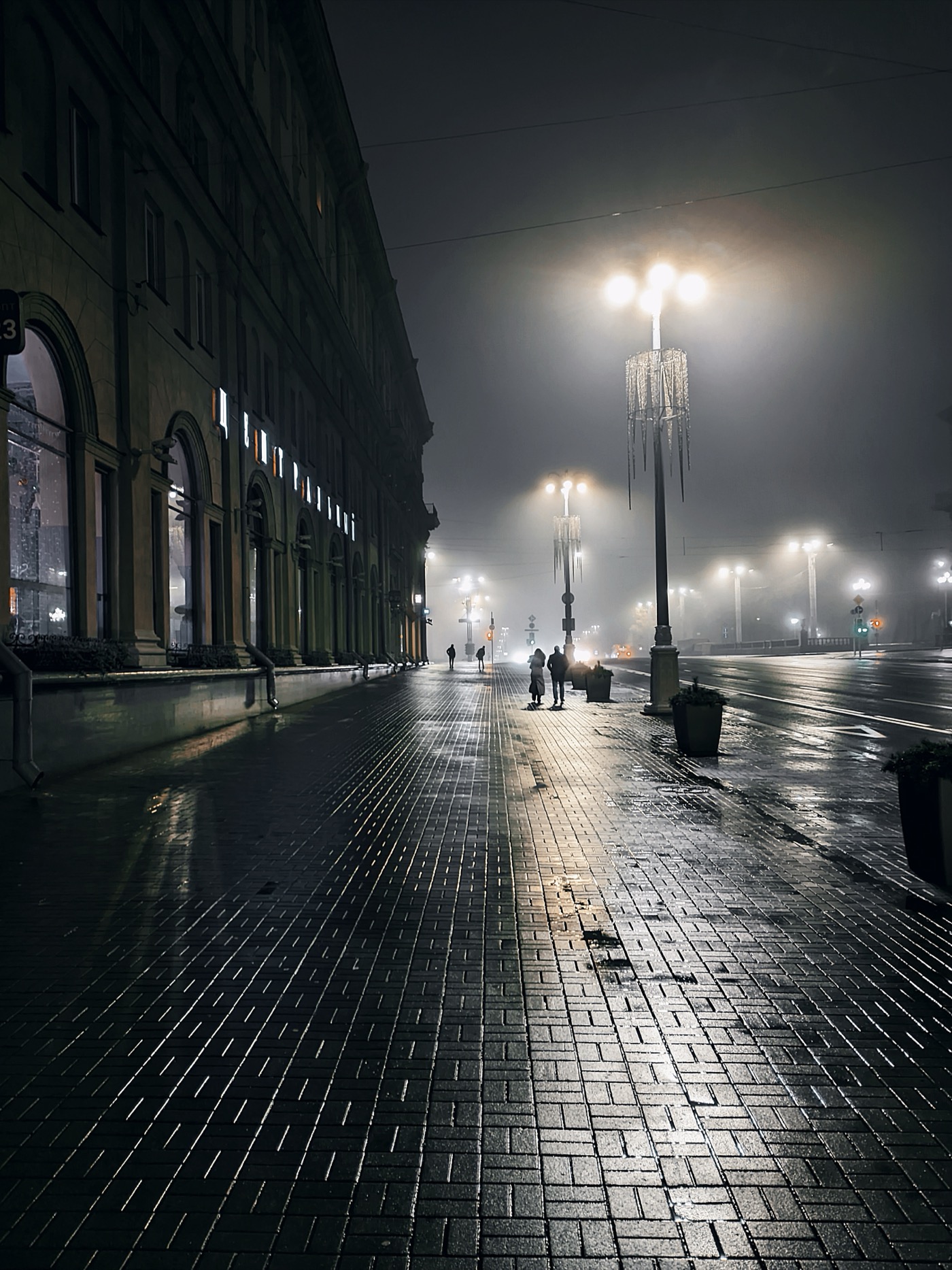 Ночной город с фонарями и туманом, по тротуару идут люди, отражения на мокрой плитке.