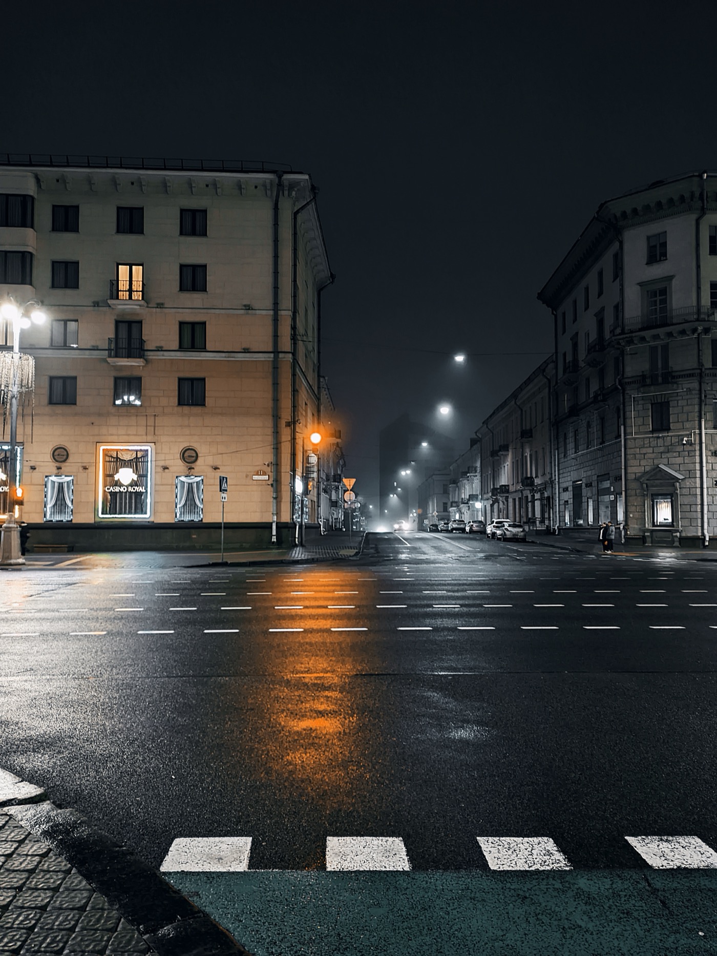 Ночной городской пейзаж с мокрой улицей, освещенной фонарями, и зданиями по обеим сторонам.