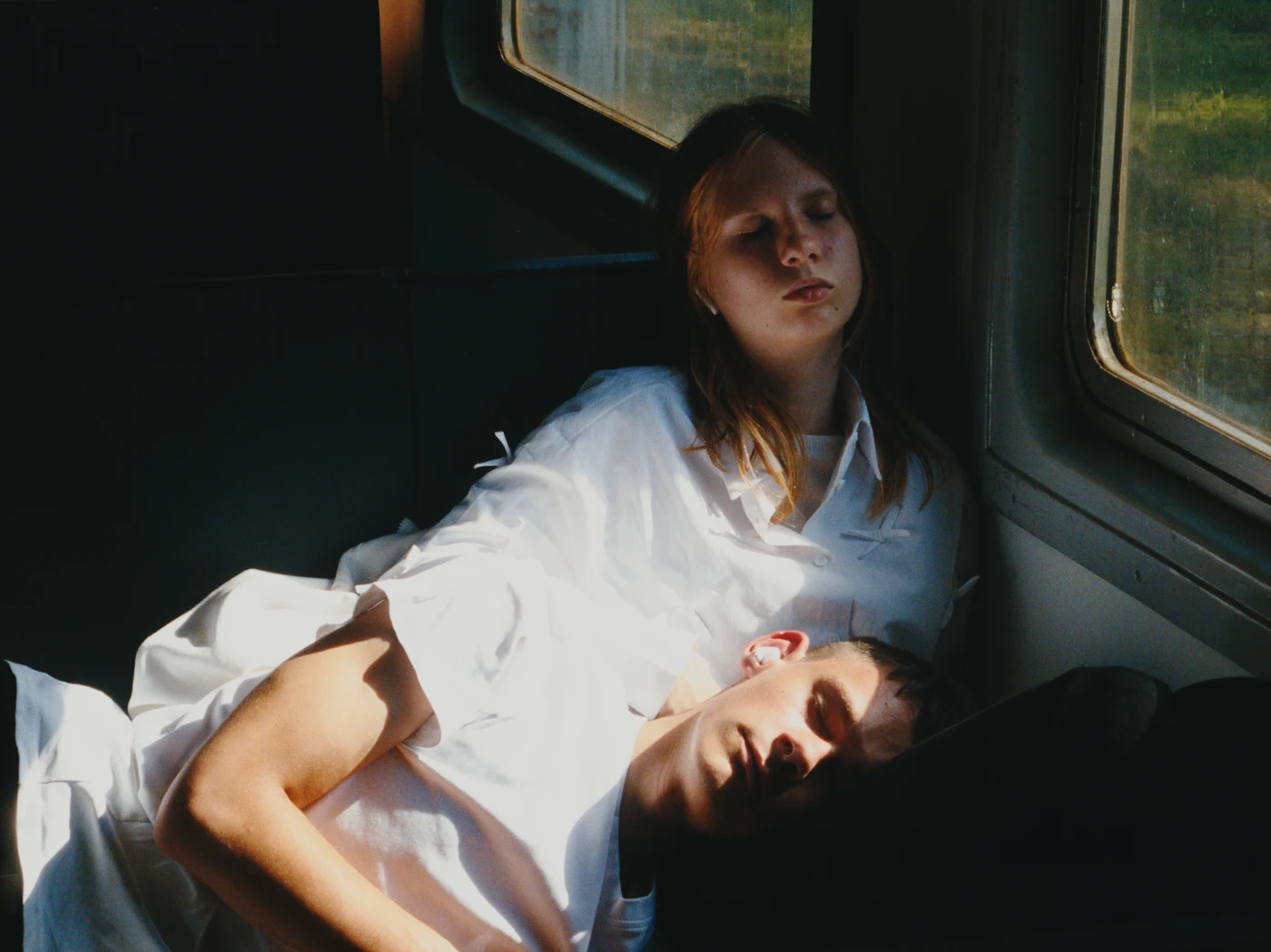 Two people dressed in white sleep on train seats, one resting their head on the other’s shoulder.