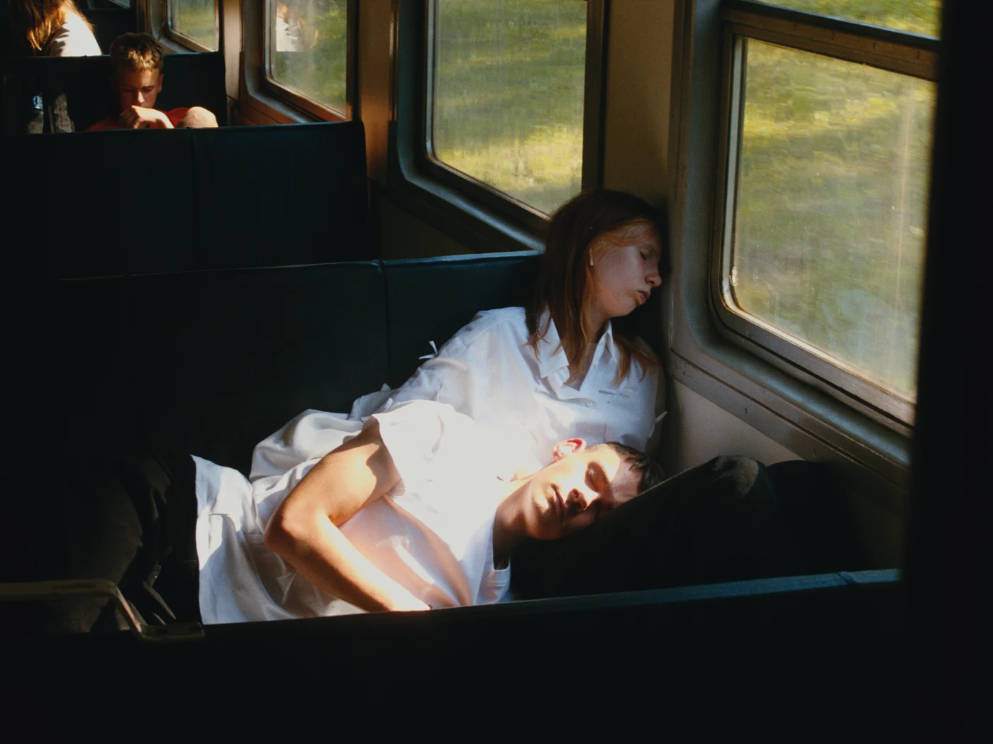 A young couple sleeps on train seats, their heads leaning toward each other. A green landscape is visible through the window.