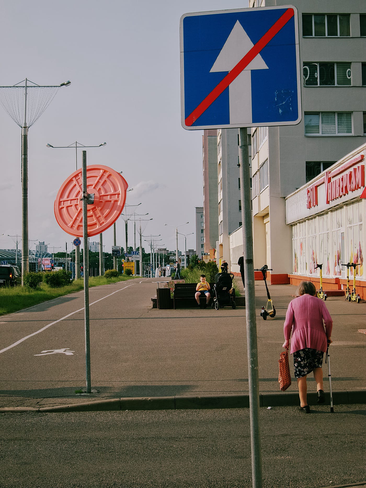 Пожилая женщина проходит возле дорожного знака на фоне вечернего города