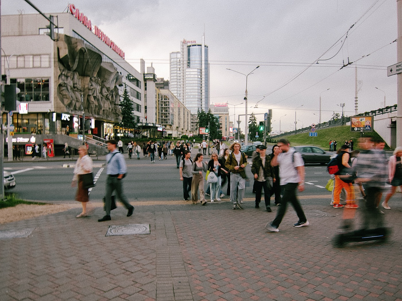 Люди переходят улицу у торгового центра, на фоне здания и небоскрёбов, вечернее время.