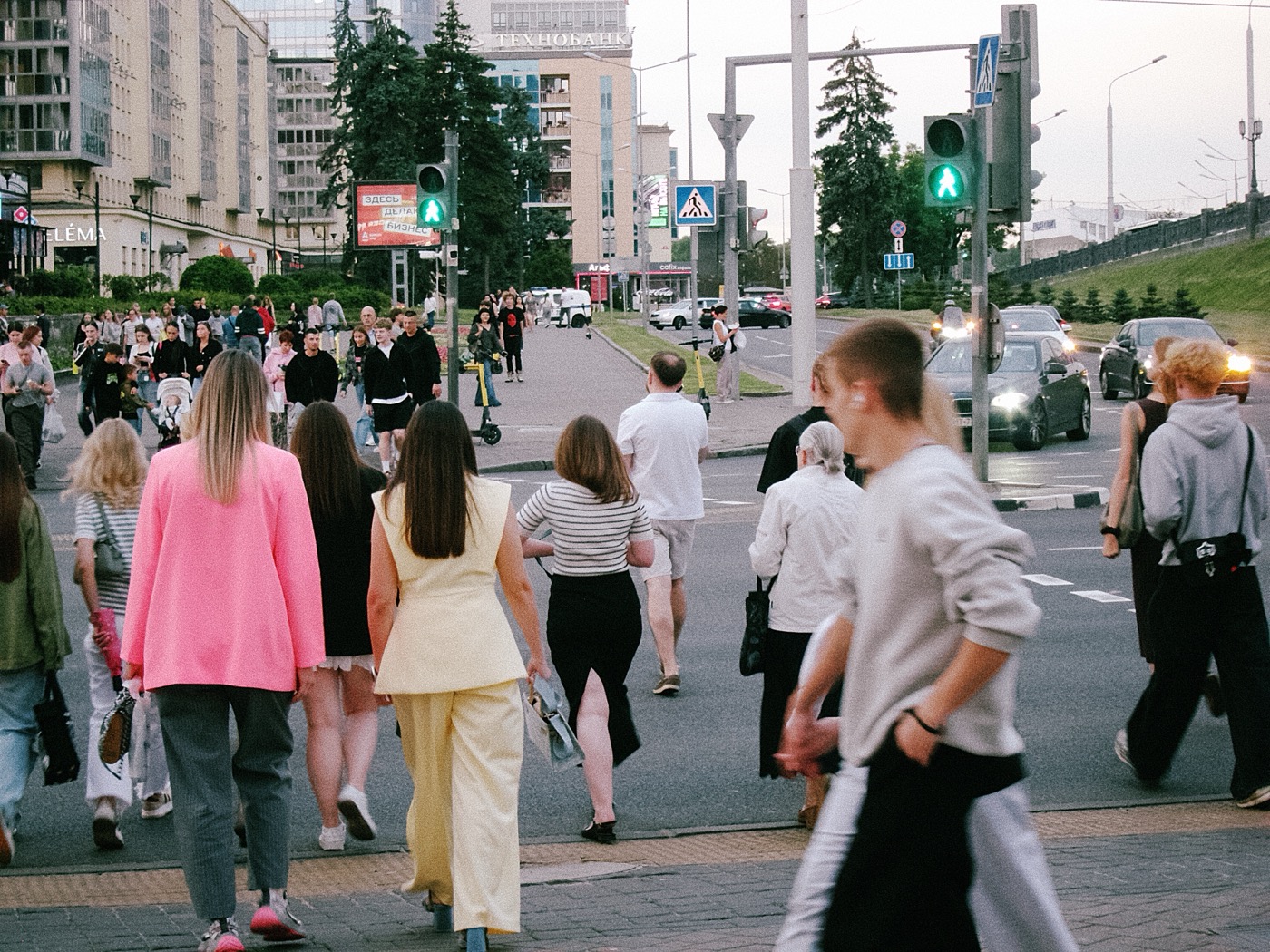 Люди переходят улицу на светофоре, вдалеке видны здания и автомобили.