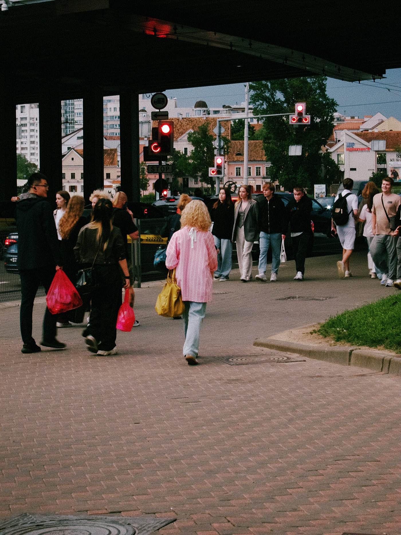 Группа людей на улице, некоторые с пакетами, переходят дорогу на красный свет светофора.