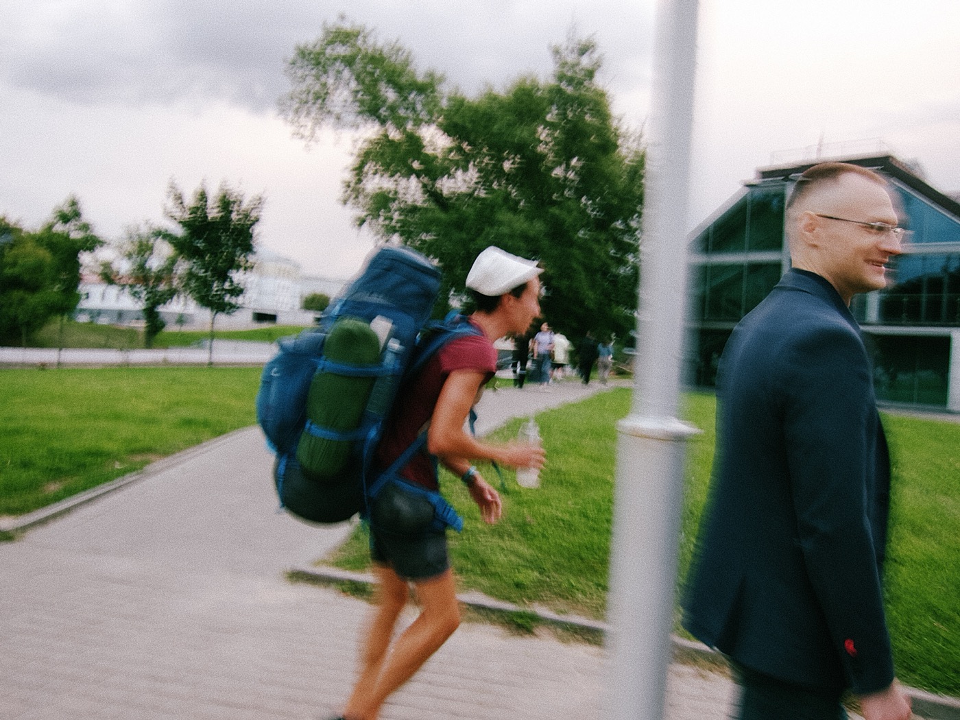 Мужчина с рюкзаком идет по тропинке, рядом улыбающийся человек в костюме. На заднем плане зелёные деревья и здания.