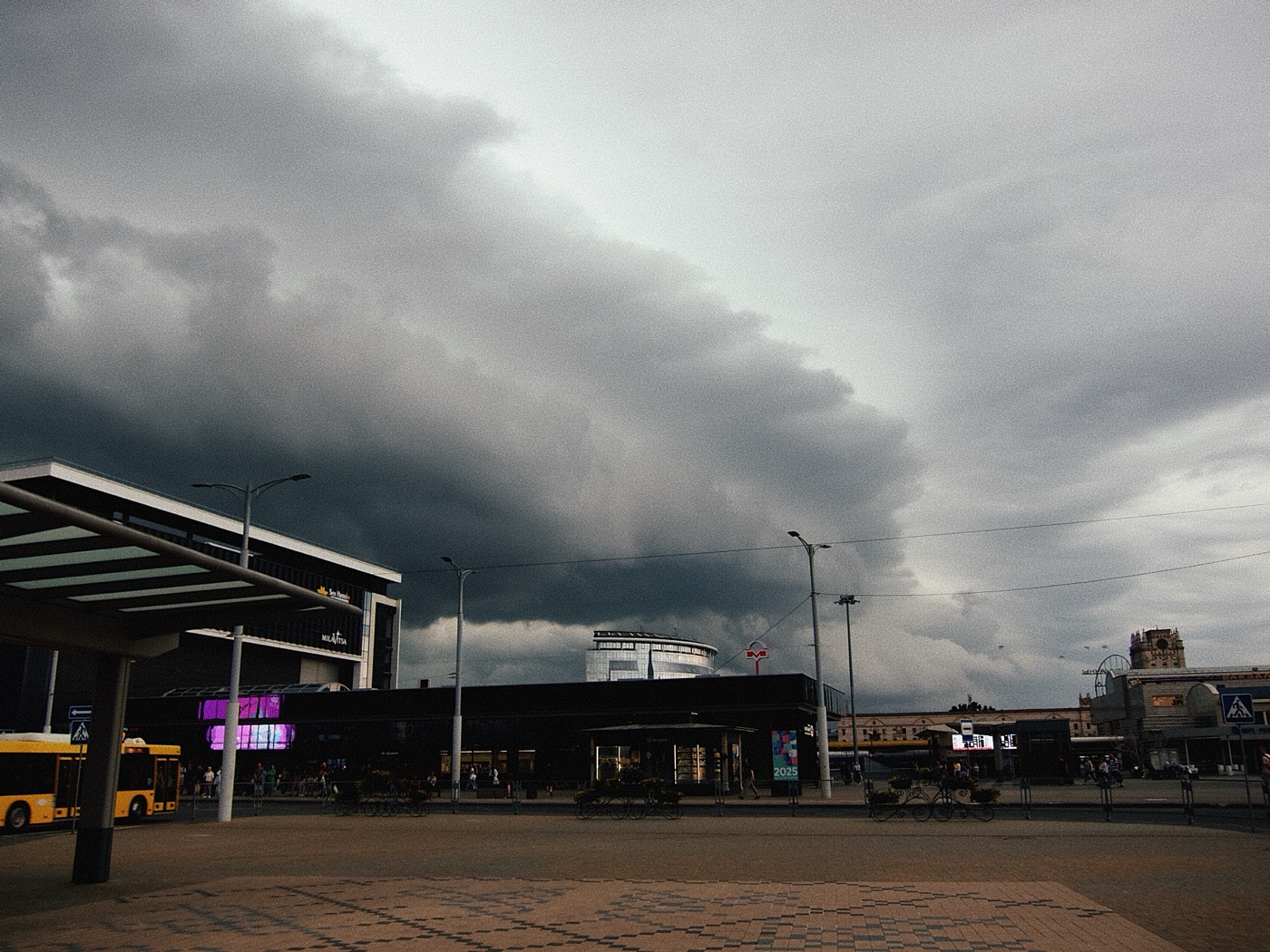 Темное облачное небо над городской площадью, здания и автобусная остановка на переднем плане.