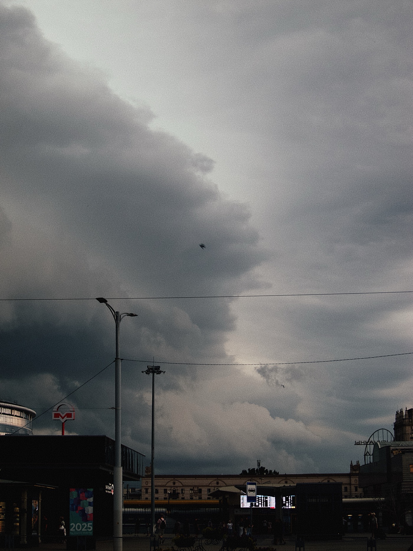 Темное облачное небо с небольшими просветами, на переднем плане городская улица с людьми и рекламными щитами.