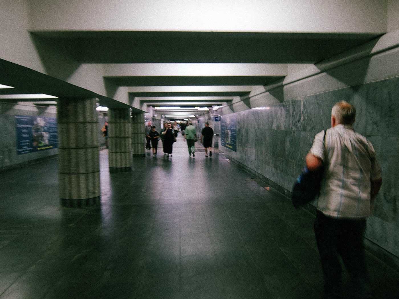 Люди идут по длинному коридору в метро, стены отделаны серым мрамором.