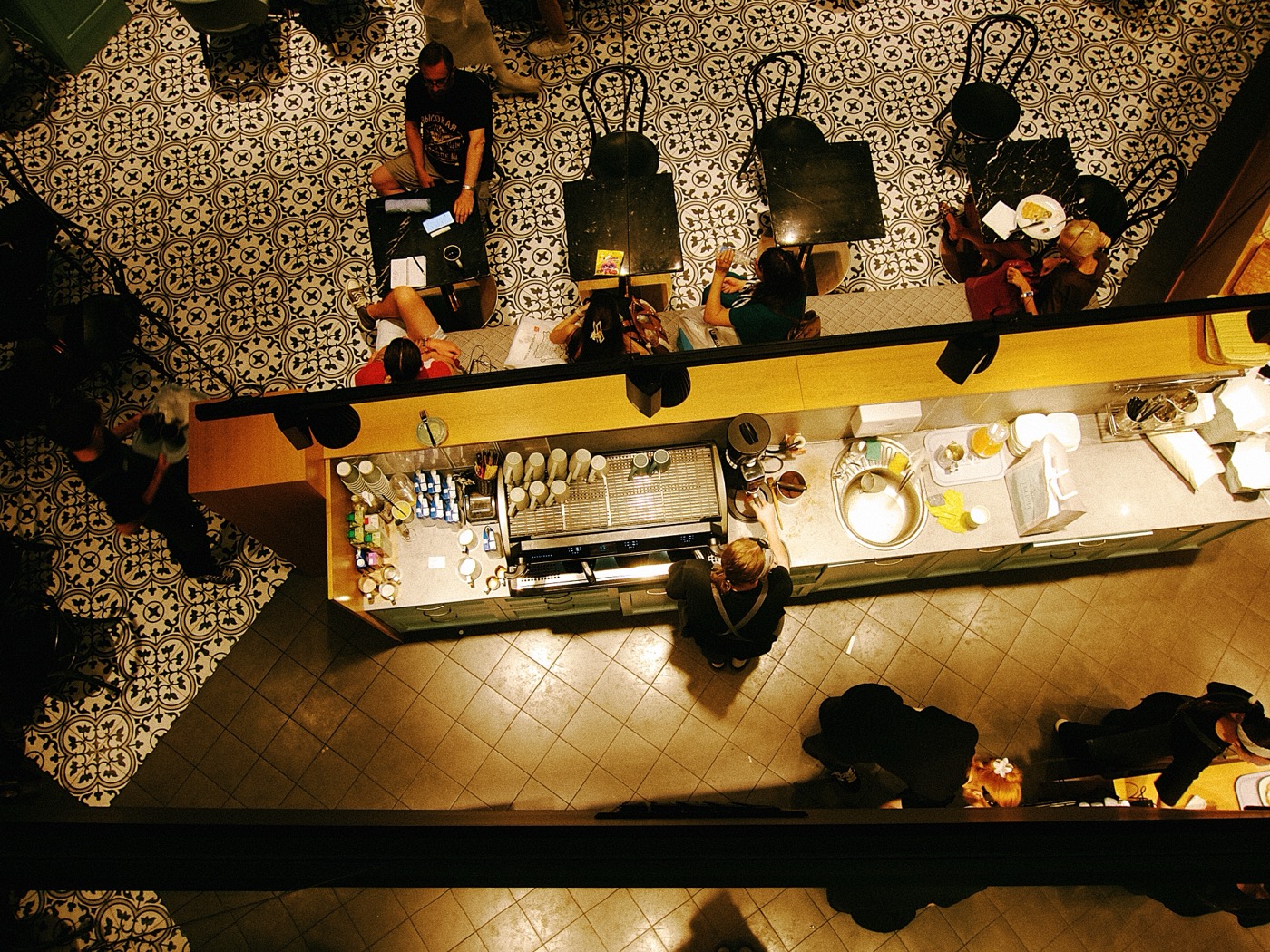 Вид сверху на кафе с посетителями, столами и барной стойкой. На полу узорчатая плитка.