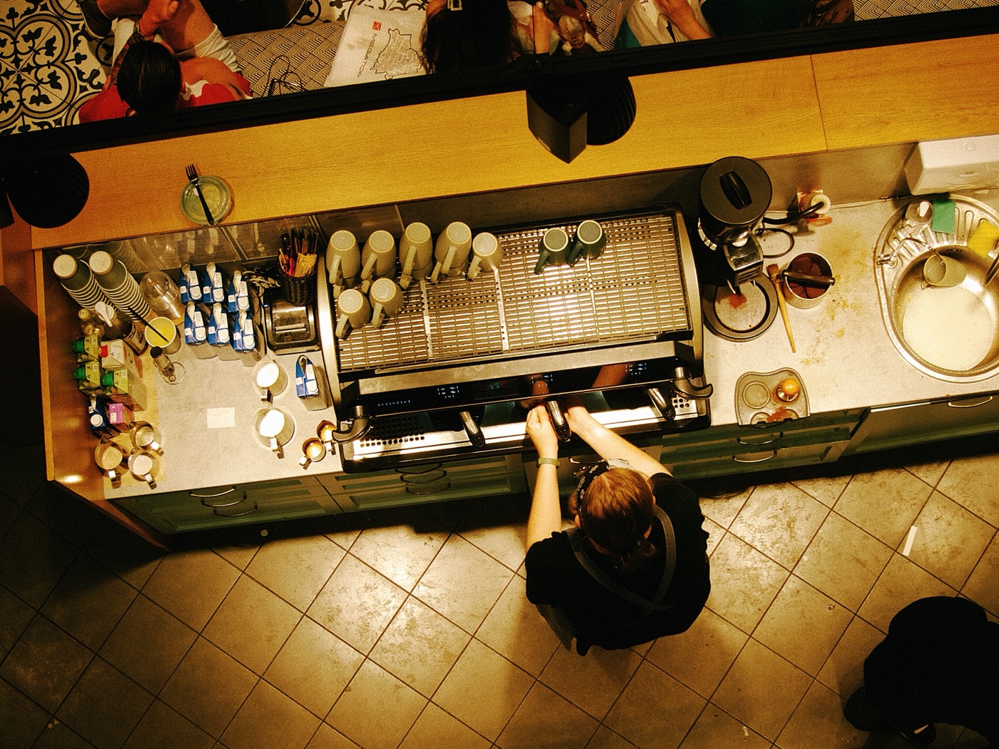 Бариста готовит кофе на кофемашине, вокруг расположены чашки и ингредиенты. Сверху виден интерьер кафе.