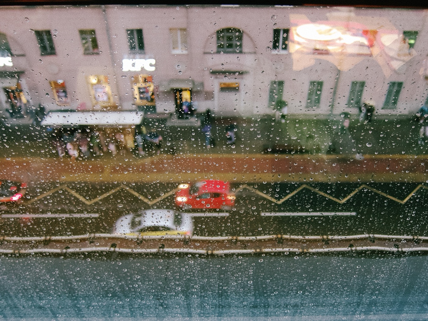 Дождливый день, капли на стекле, вид на улицу с проезжающими машинами и людьми.