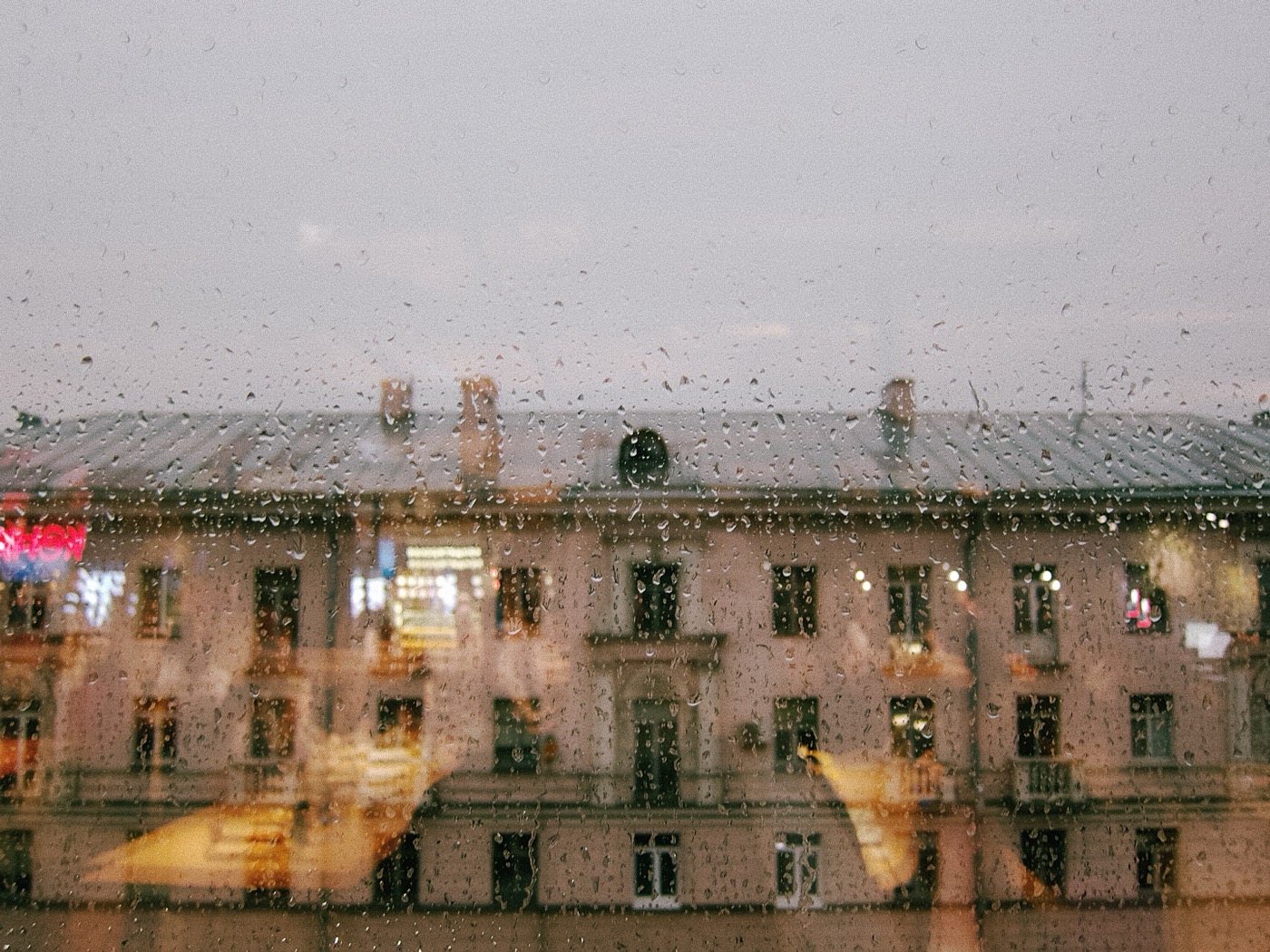 Дождливый день, капли на стекле, вид на здание с окнами и крышей. Небо серое, атмосфера спокойная.