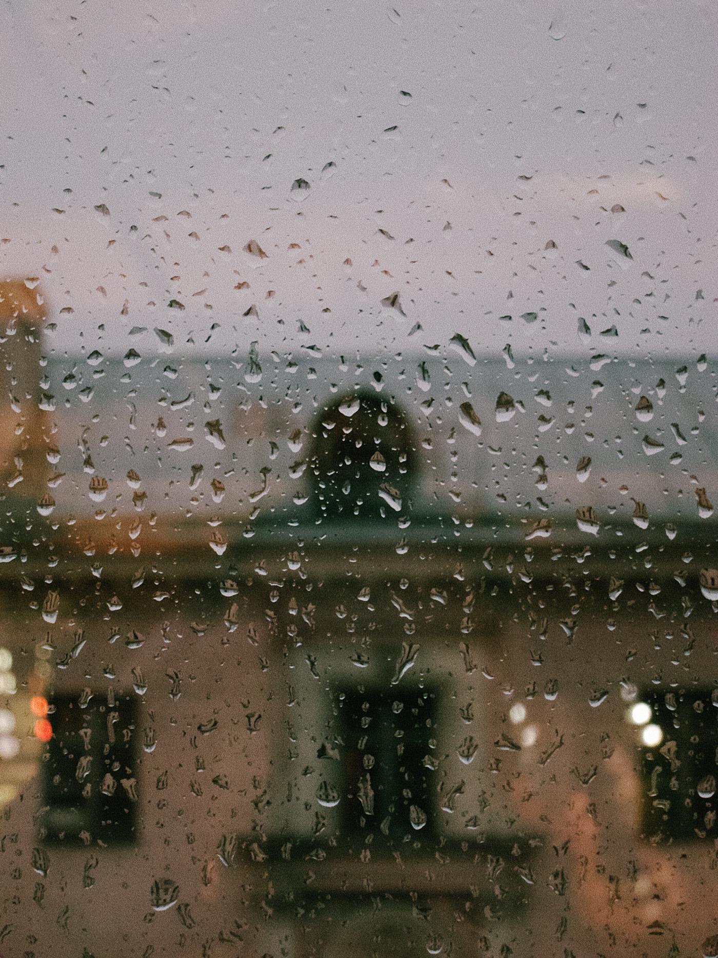 Капли дождя на стекле, за которым виден городской пейзаж в сумерках.