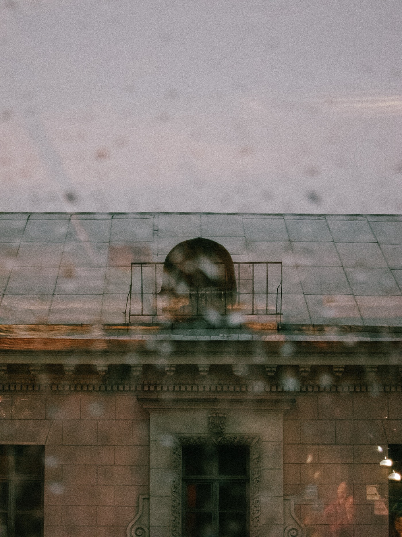 Вид на здание с крышей и балконом через дождливое окно. Небо затянуто облаками.