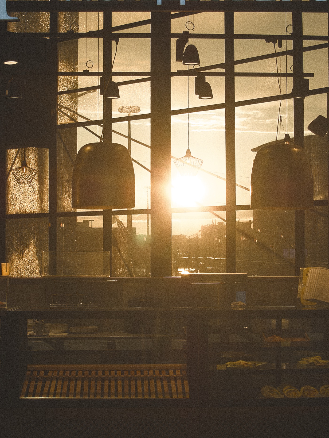 Солнечный свет проникает через большие окна кафе, создавая теплую атмосферу. На переднем плане видны светильники.