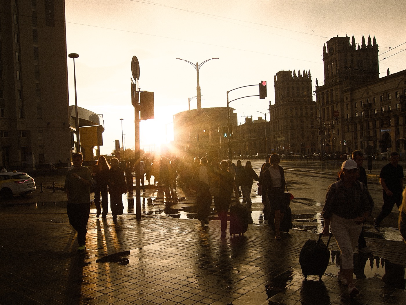 Силуэты людей на улице на фоне заката, отражения на мокрой плитке. Светофор и здания вдалеке.