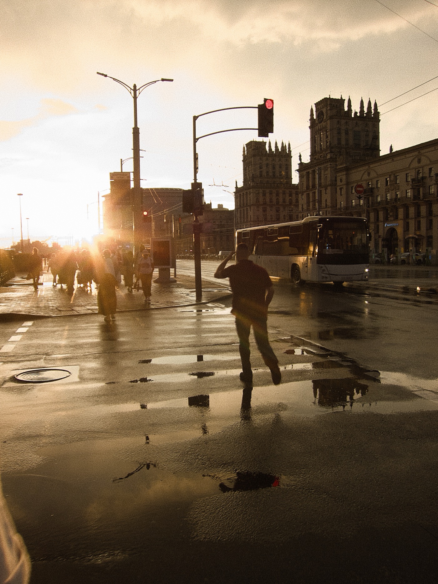 Силуэт человека на мокрой улице, яркие огни светофора и отражения в лужах на фоне зданий.