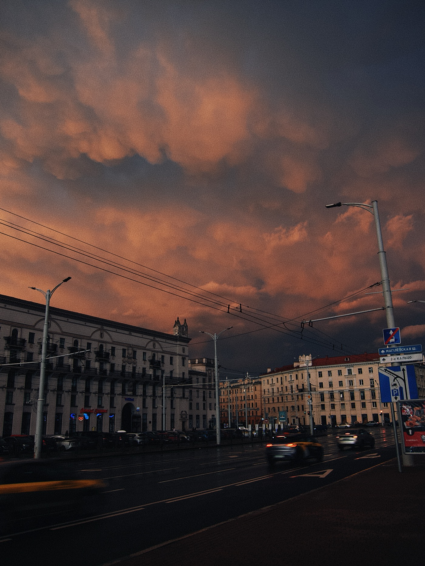 Закатное небо с облаками над городской улицей, автомобили движутся мимо зданий.