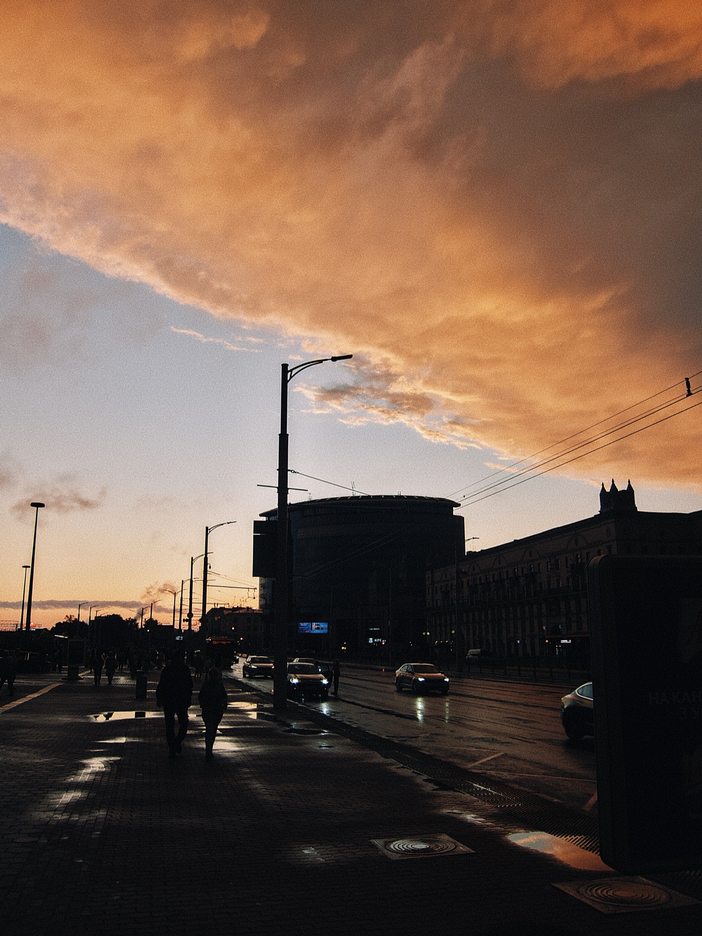 Закат с яркими облаками над городом, silhouettes людей и автомобилей на улице.