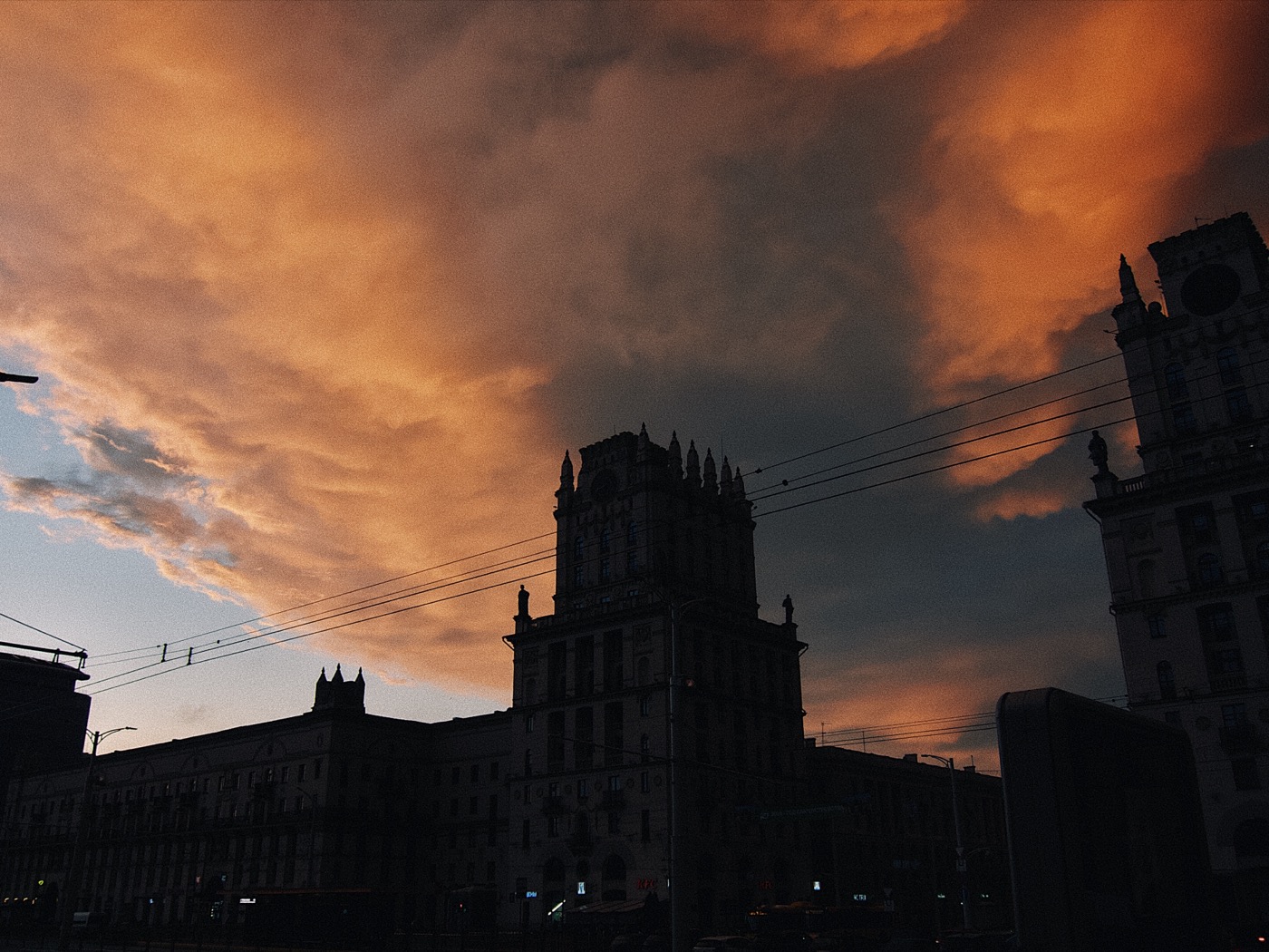 Закат над городом, облака окрашены в оранжевые и фиолетовые оттенки, silhouetted здания на переднем плане.