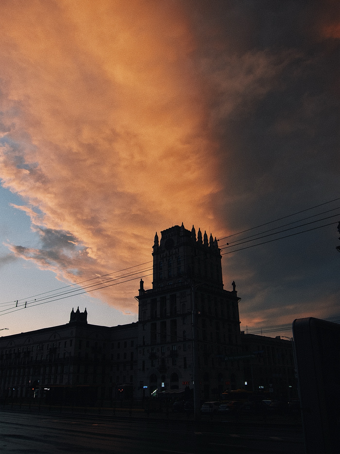 Закат над городом, облака окрашены в оранжевые и фиолетовые оттенки, силуэт здания на переднем плане.