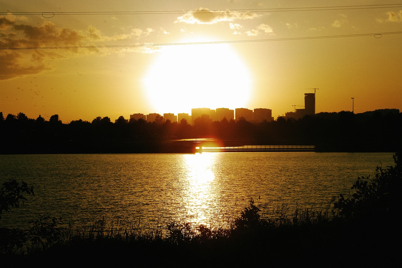 Закат над городом, солнце отражается в воде, silhouetted здания на фоне.