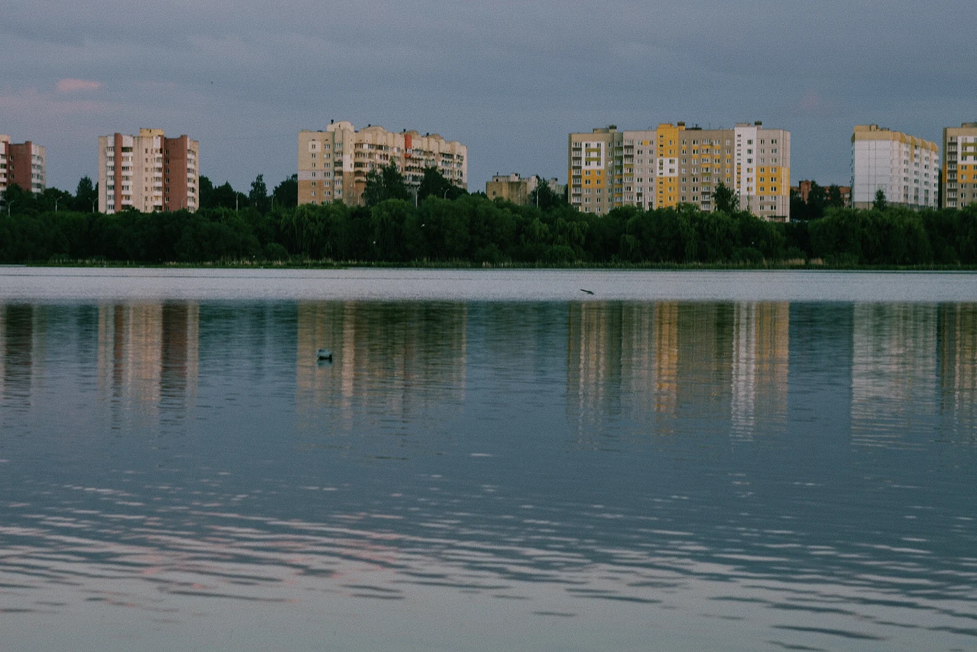 Городские здания отражаются в спокойной воде озера под серым небом.