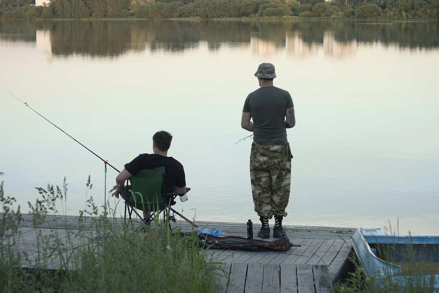 Two fishermen on the shore of a lake; one sits on a chair, the other stands with a fishing rod. Calm water and greenery all around.