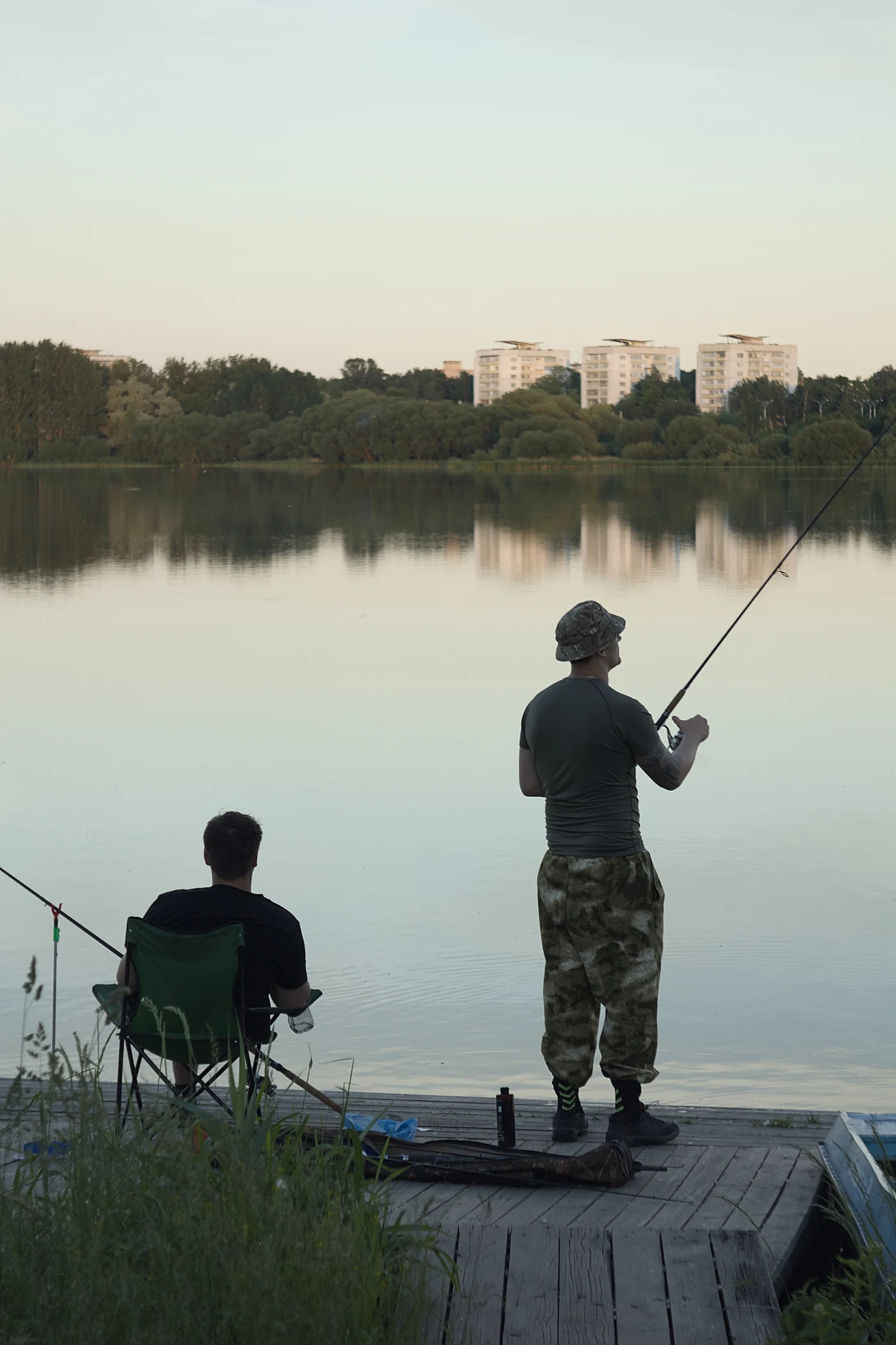 Two fishermen on the shore of a lake, one of them fishing, with apartment buildings visible in the background.