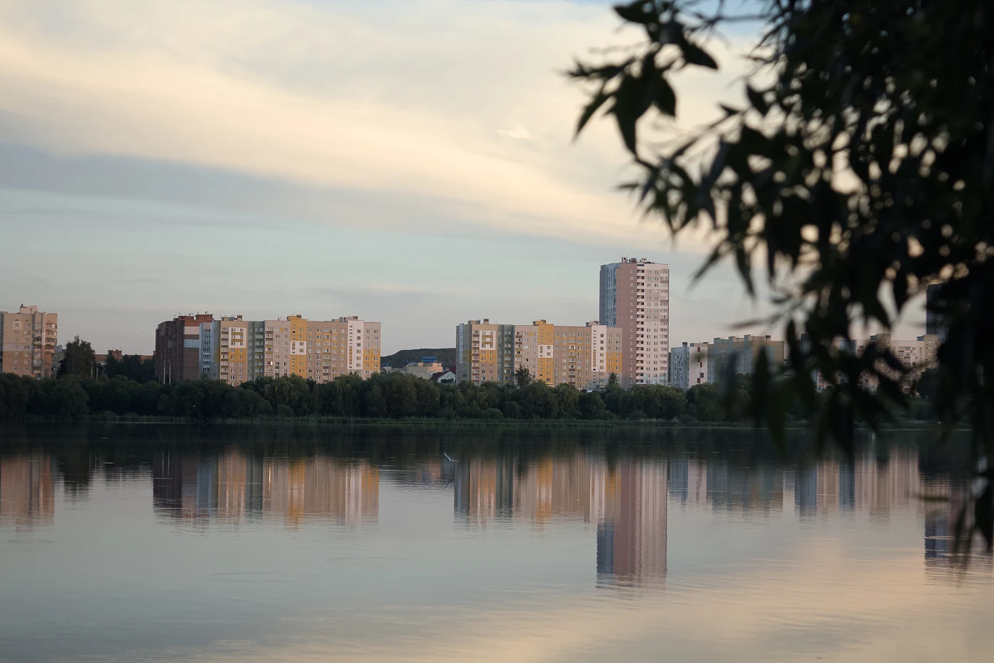 City buildings are reflected in calm water, with a cloudy sky and green trees in the background.