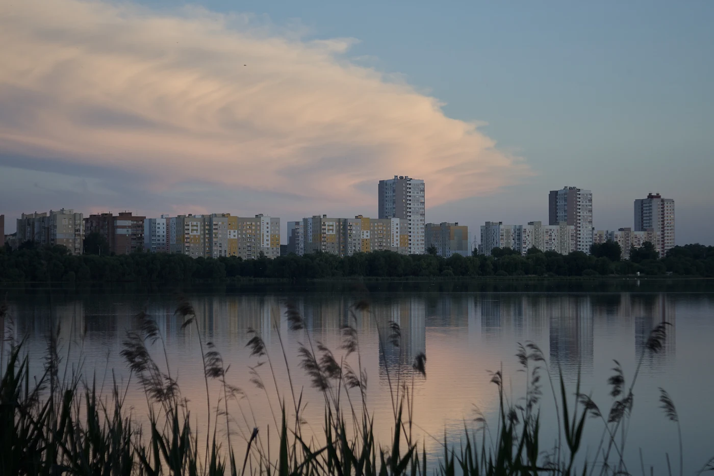 City buildings beside a calm lake and sunset clouds. Vegetation in the foreground.