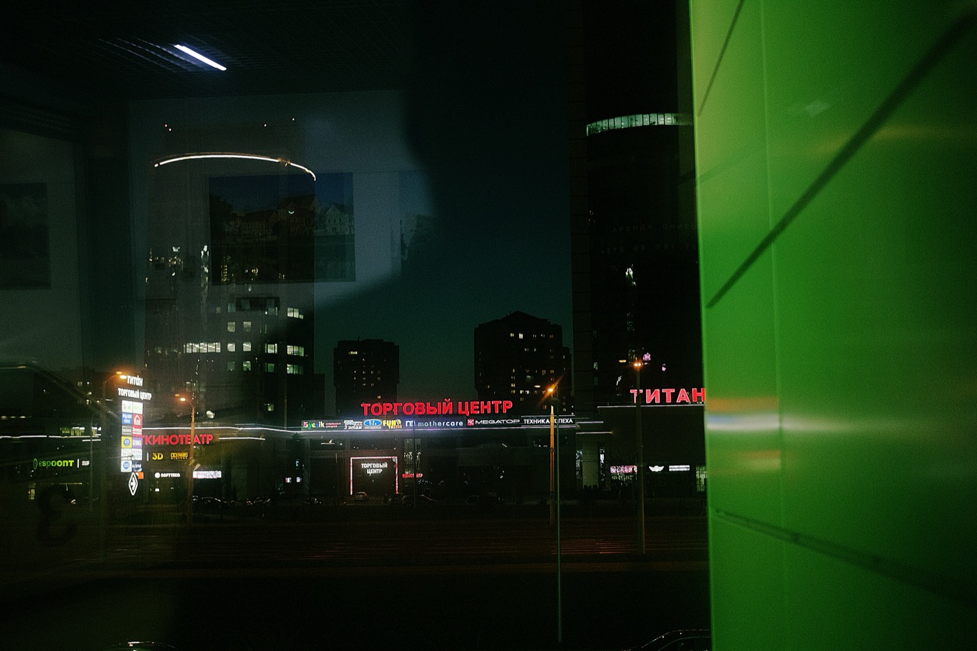 Ночной городской пейзаж с отражениями и вывеской «Торговый центр». Зелёная стена на переднем плане.