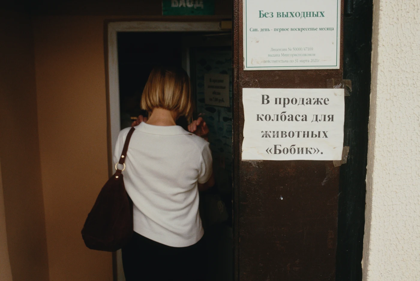 Женщина с сумкой заходит в дверь, на которой вывеска: «В продаже колбасы для животных »Бобик«».