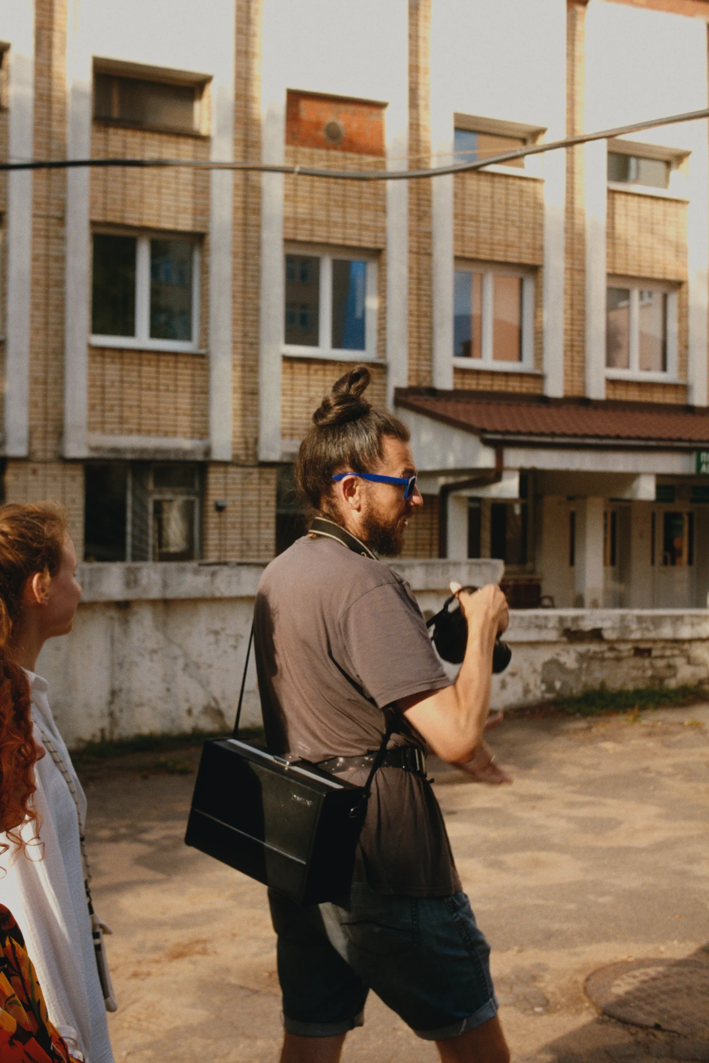 Человек с фотоаппаратом и рюкзаком на фоне многоквартирного дома, рядом девушка с вьющимися волосами.