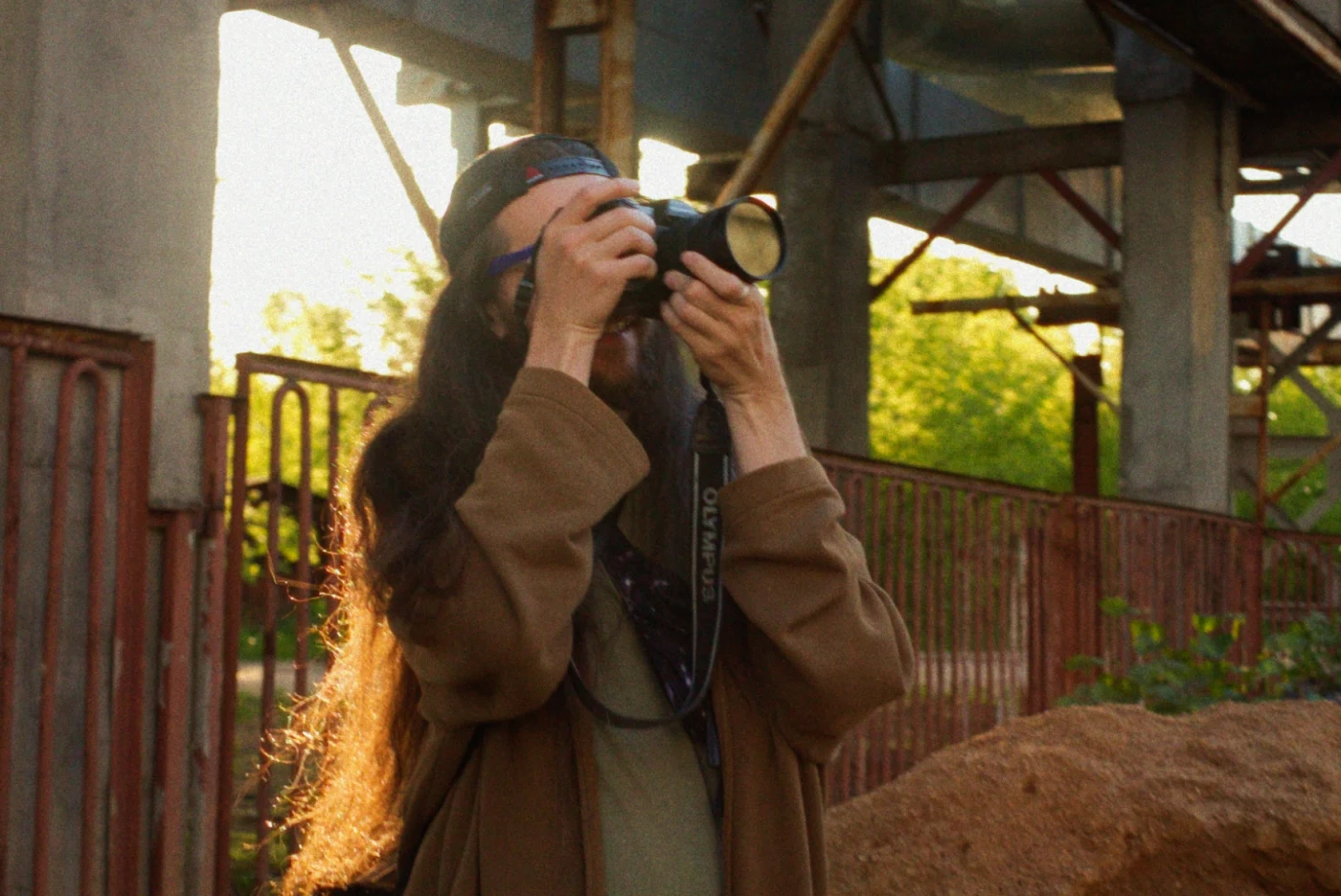 A person with long hair photographs on the street, standing against an abandoned structure and greenery.