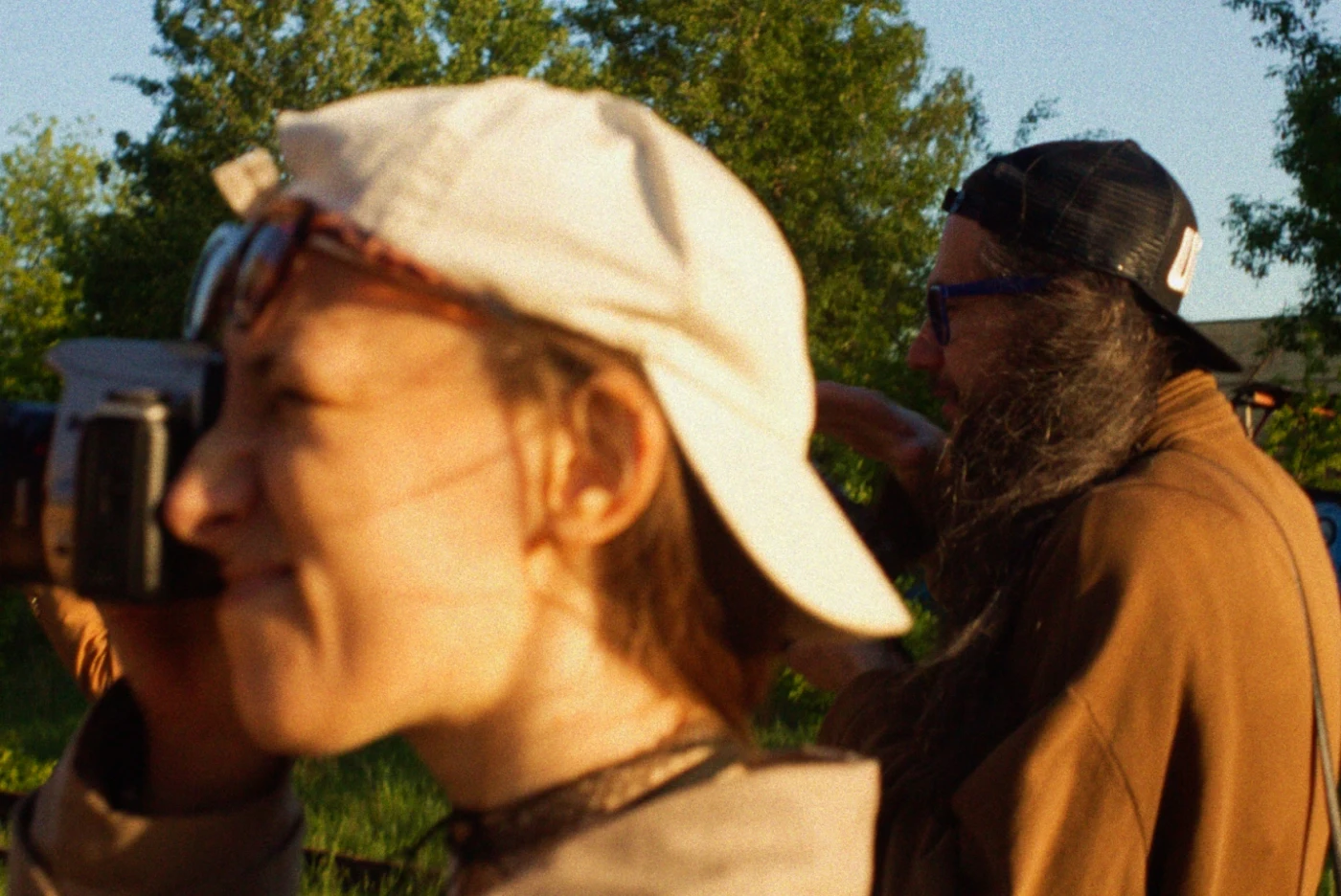 A woman with a camera, with a bearded man in the background, both outside among greenery.