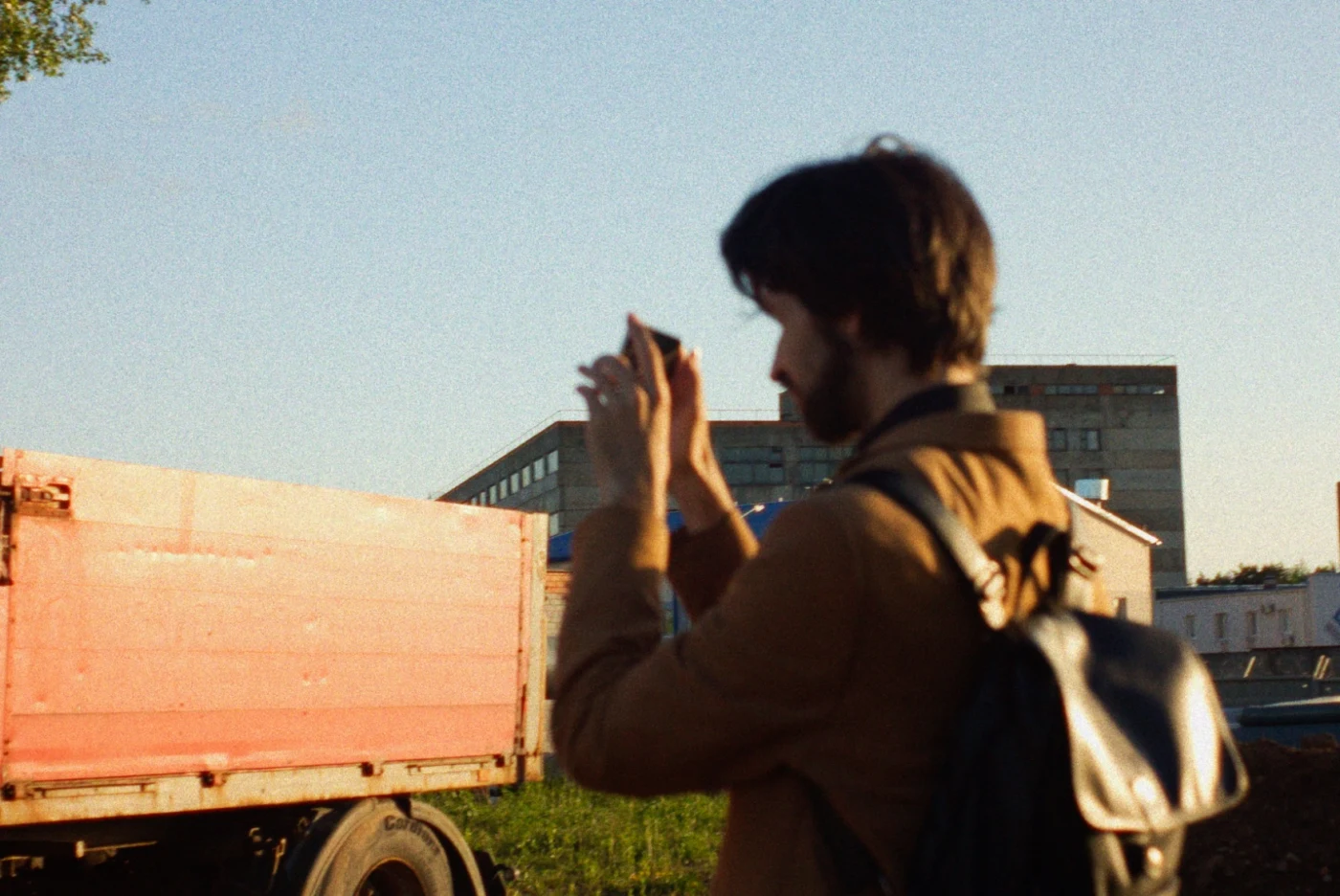 A person with a backpack photographs against a background of a truck and buildings under a clear sky.
