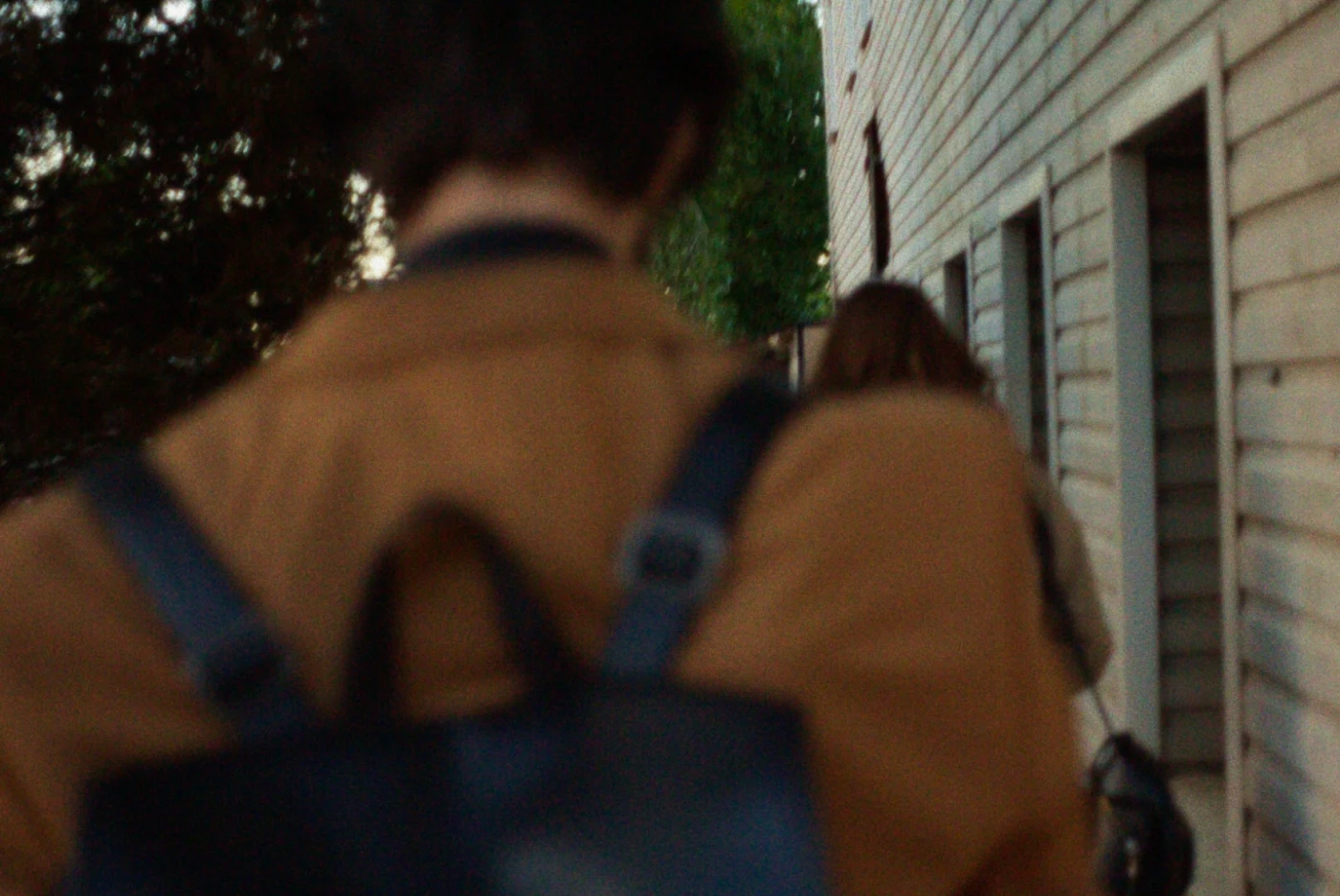 Two people walk along a wooden wall, one in a brown coat with a backpack, the other in light-colored clothing.