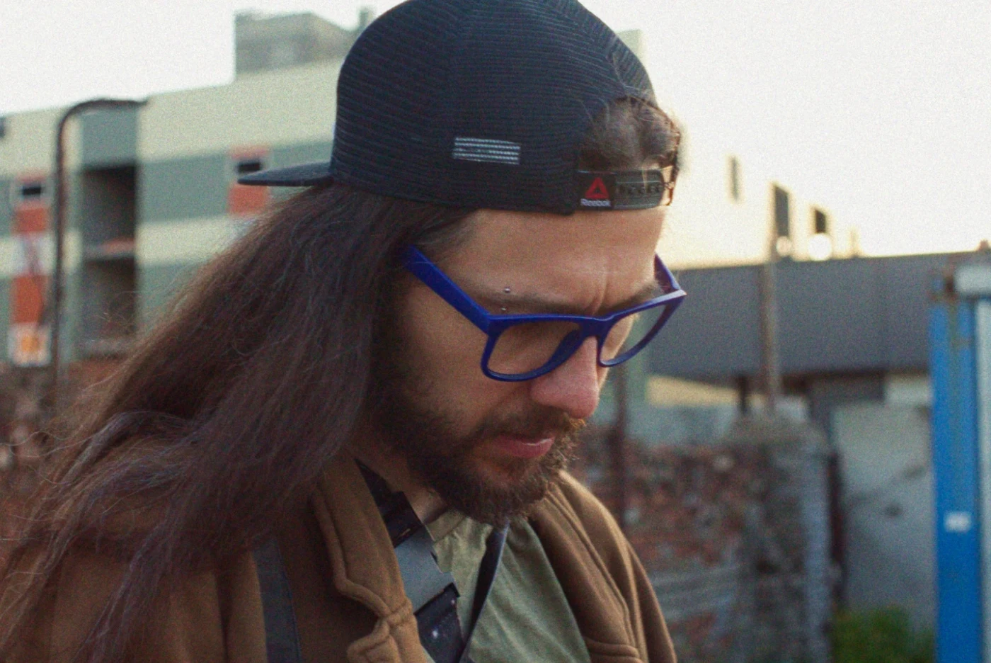 A man with long hair and blue glasses, wearing a brown jacket, looks down against an urban landscape.
