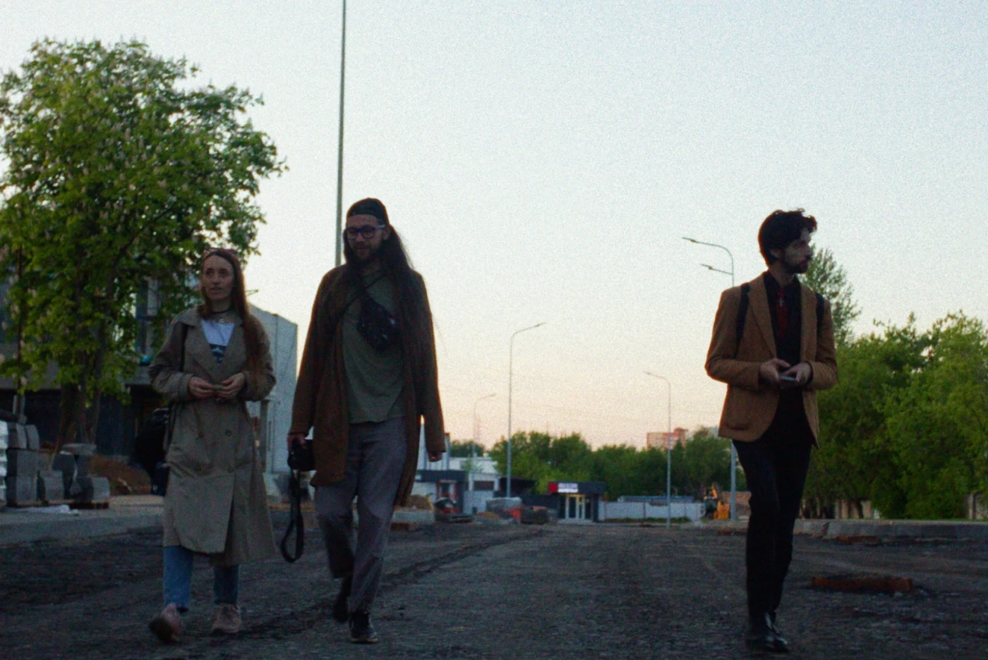 Three people walk along an empty street against an evening sky and green trees.