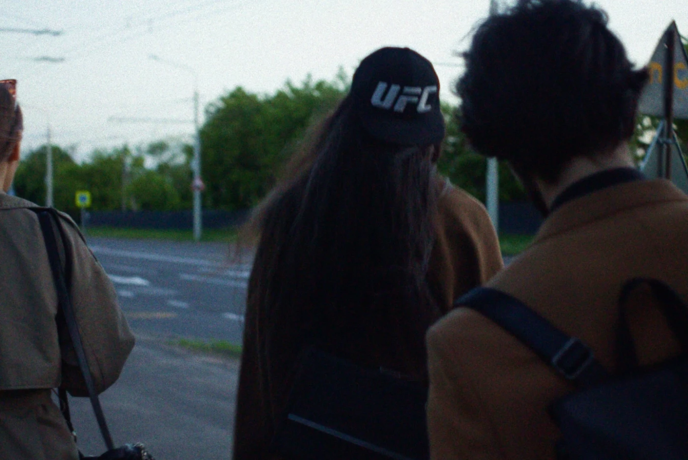 A group of people walk along the street, one of them wearing a UFC cap, with trees in the background.