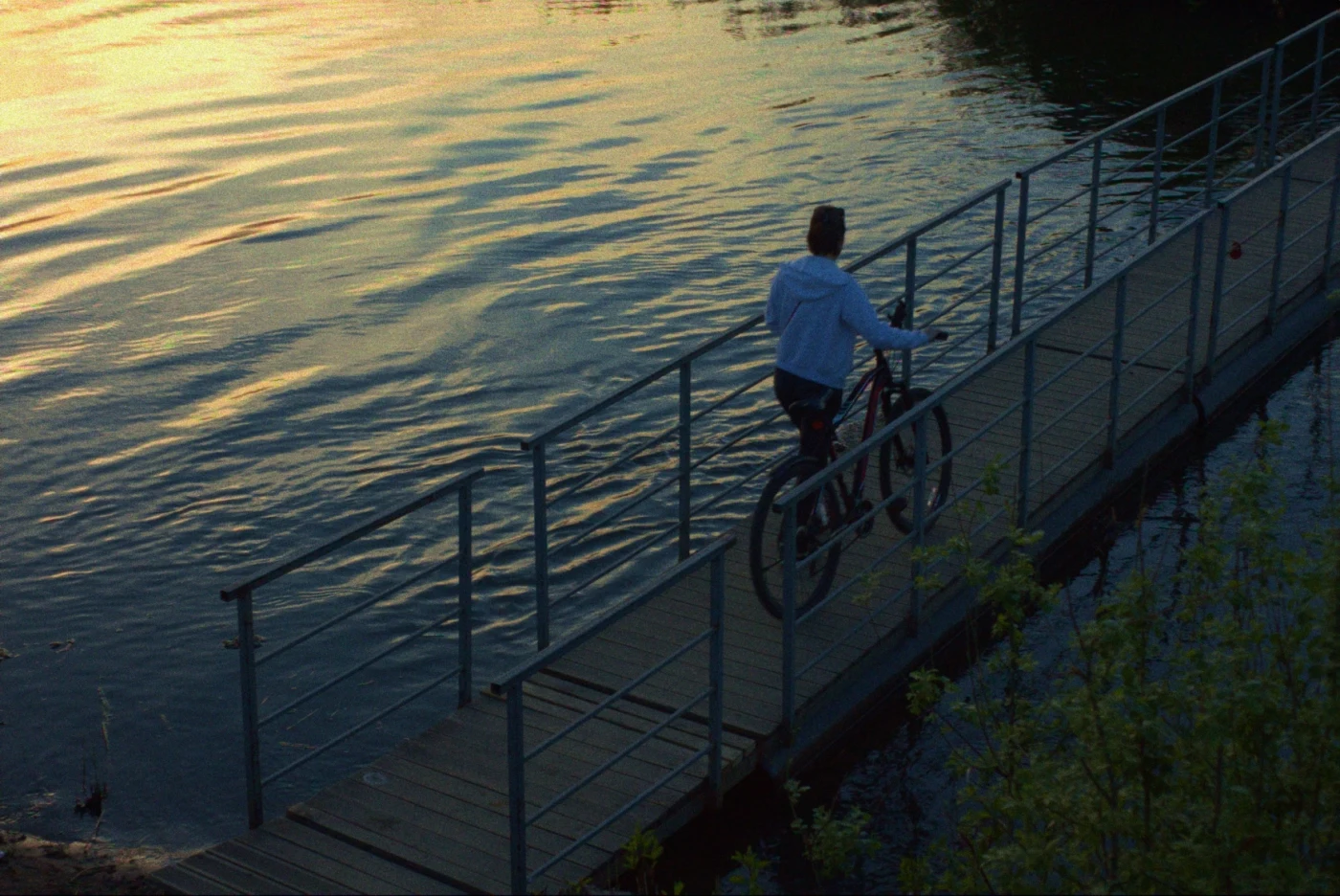 Человек на велосипеде едет по деревянному мосту над водой на фоне заката.